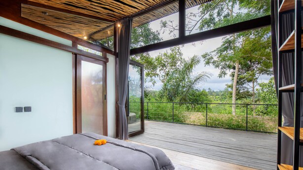 Une chambre dotée d’un grand lit, d’une porte-fenêtre coulissante donnant sur un balcon et offrant une vue sur la verdure environnante.