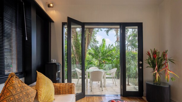 Una sala moderna con un sofá, cojines estampados, una mesa de comedor y vistas a un jardín tropical a través de puertas de cristal.