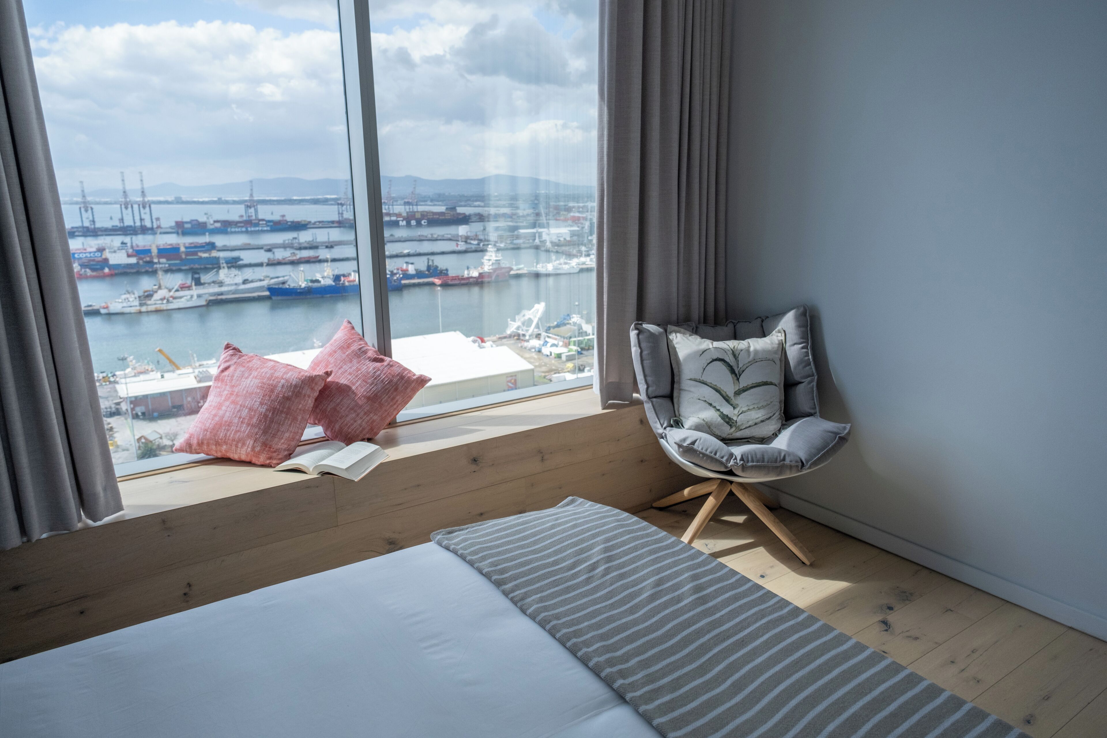 A modern hotel room with a large window overlooking a marina, a wooden floor, a bed with pillows, a grey armchair, and a striped rug.