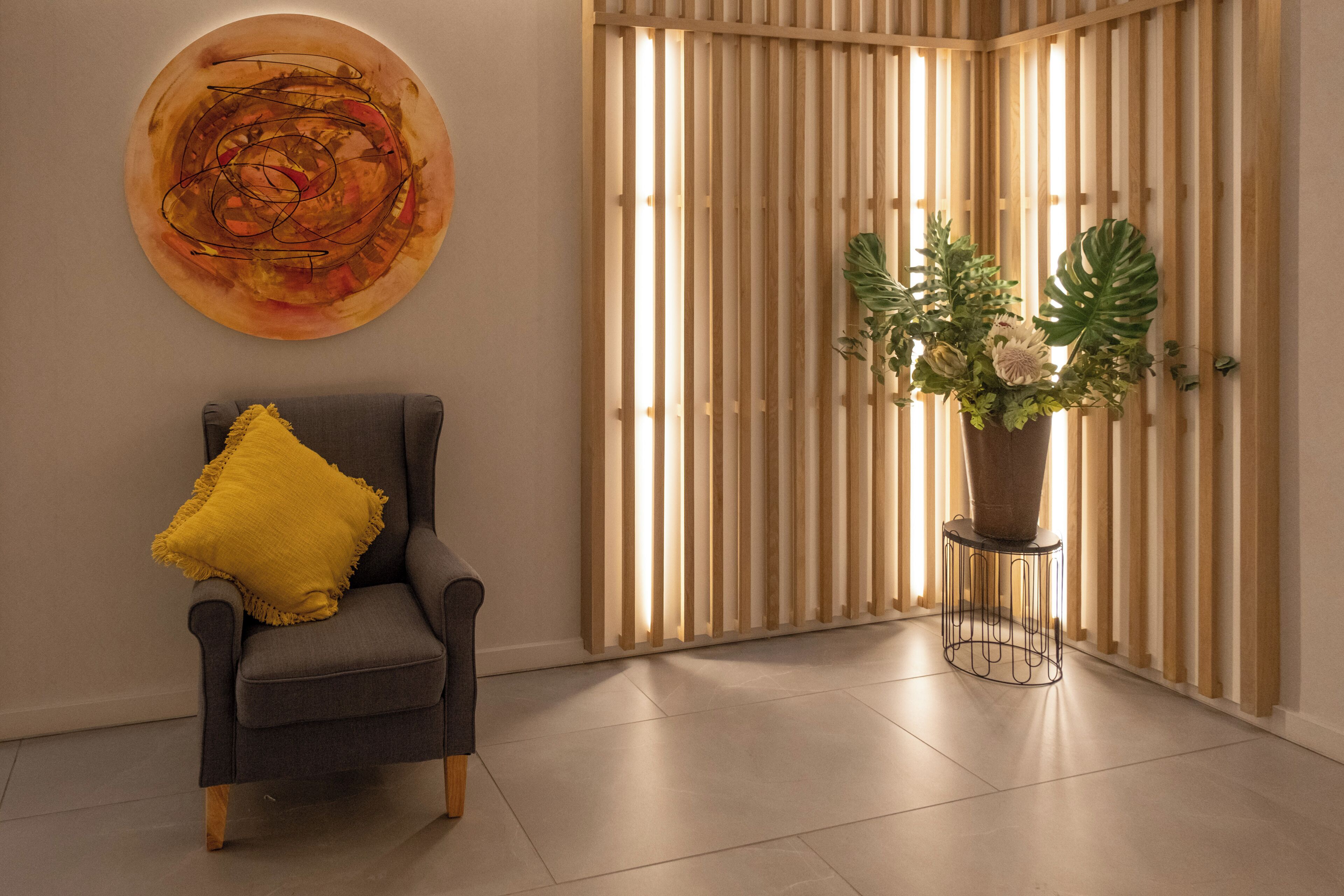 A modern room with a grey armchair, a yellow pillow, a large abstract painting, and a potted plant with white flowers.
