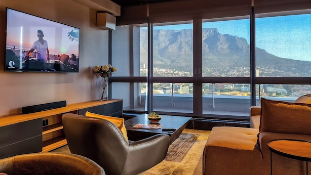 A modern living room with a large TV, comfortable seating, a coffee table, and a view of a mountain and cityscape through large windows. - The Halyard Apartments  (Cape Town)