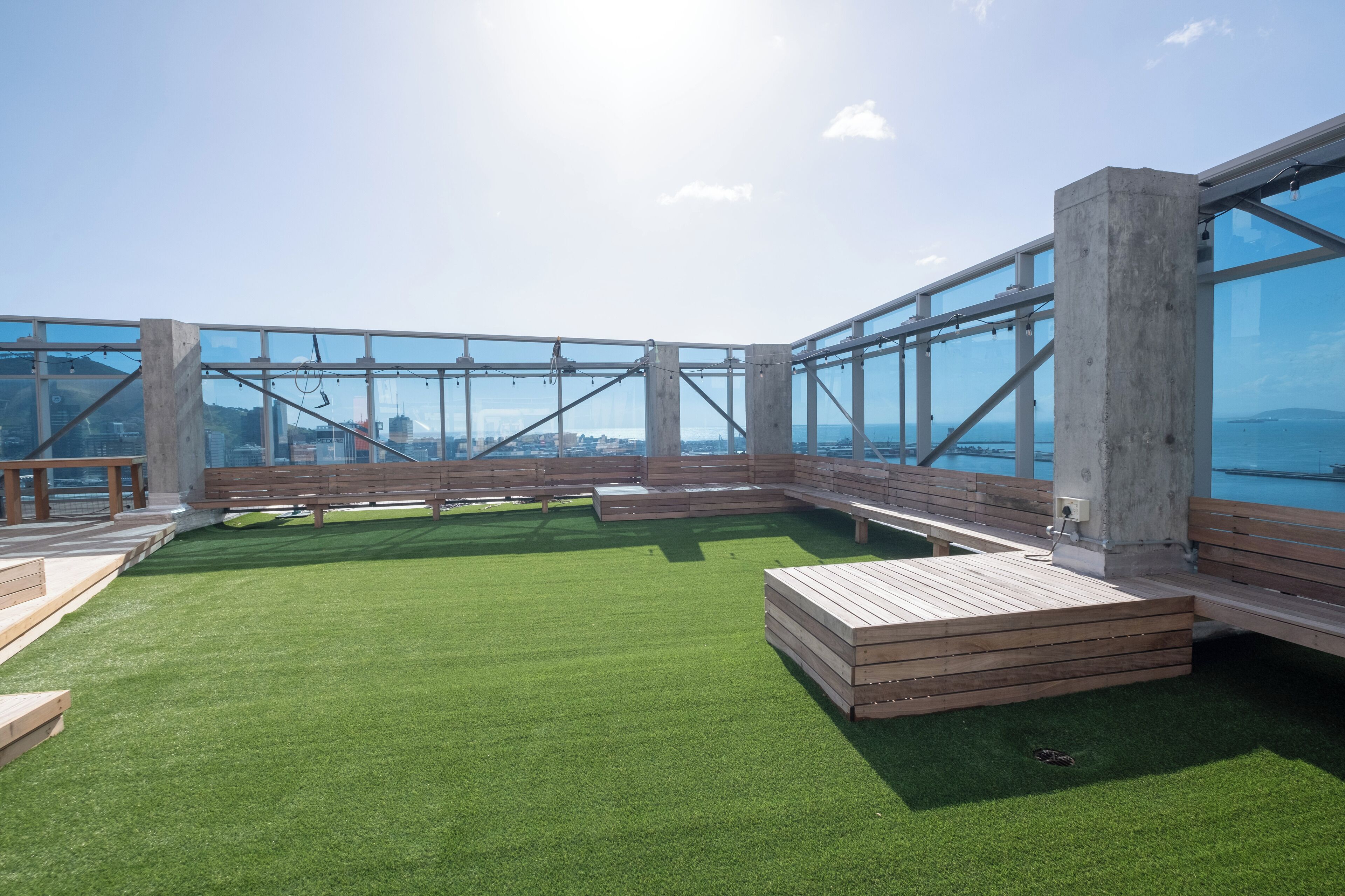 A rooftop terrace with a green lawn, wooden benches, and a glass railing overlooking a cityscape.
