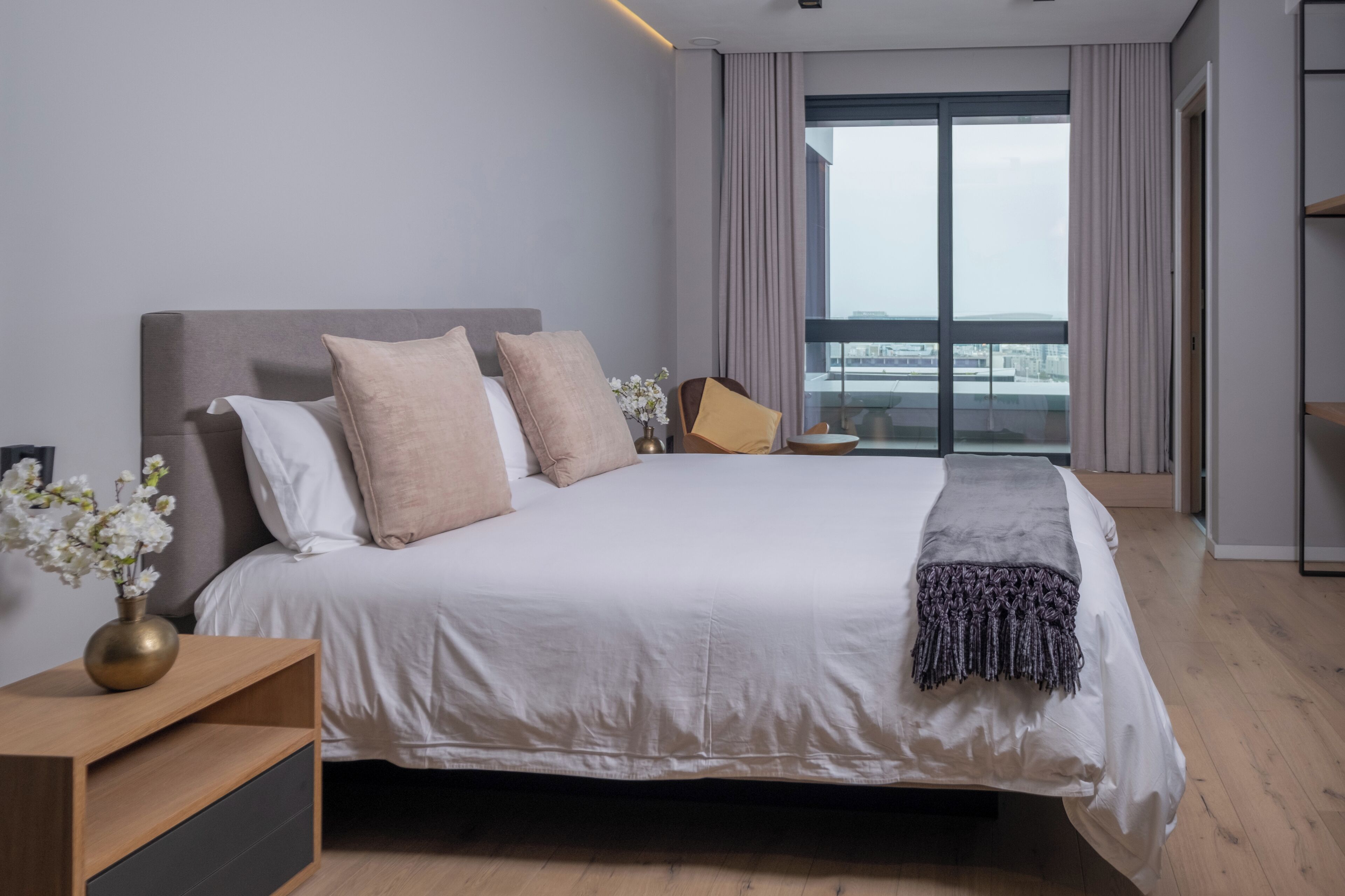 A modern hotel room with a large bed, a nightstand, a vase with flowers, and a view of the city through the window.
