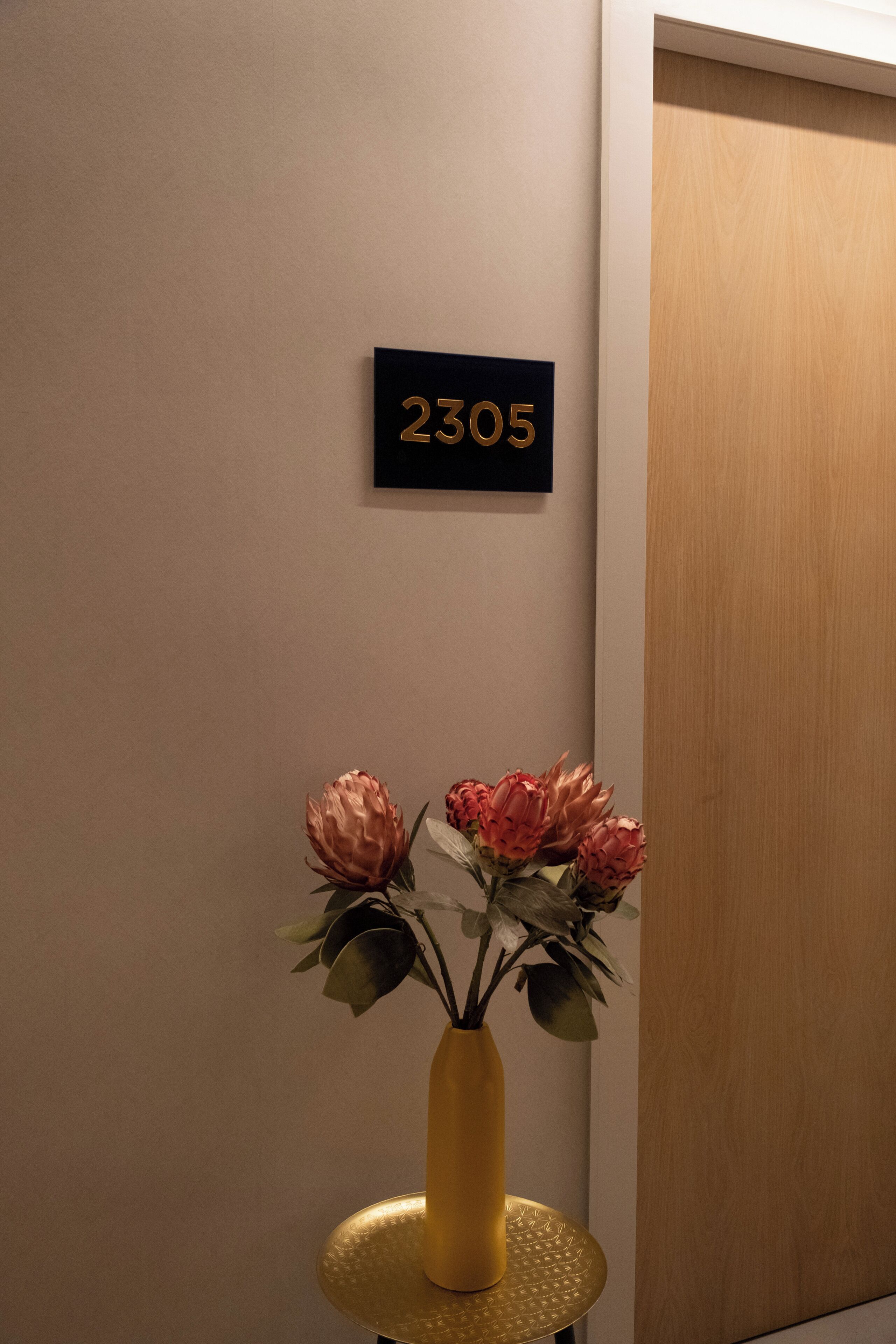 Une pièce dont le mur porte le numéro « 2305 », un vase rempli de fleurs posé sur une table ronde, et une porte en bois.