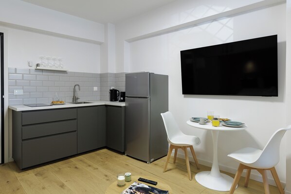 A modern kitchen with grey cabinets, a stainless steel refrigerator, a white countertop, and a dining area with two white chairs and a small table. - Lemonaki Boutique Suites (Athens)