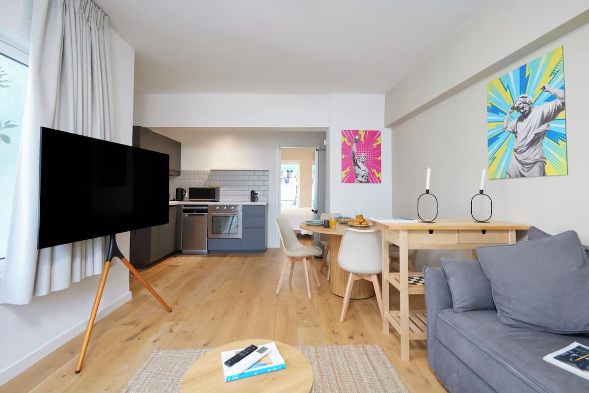 Ein modernes Wohnzimmer mit Flachbildfernseher, einem grauen Sofa, einem Holzesstisch und einer Küche im Hintergrund.