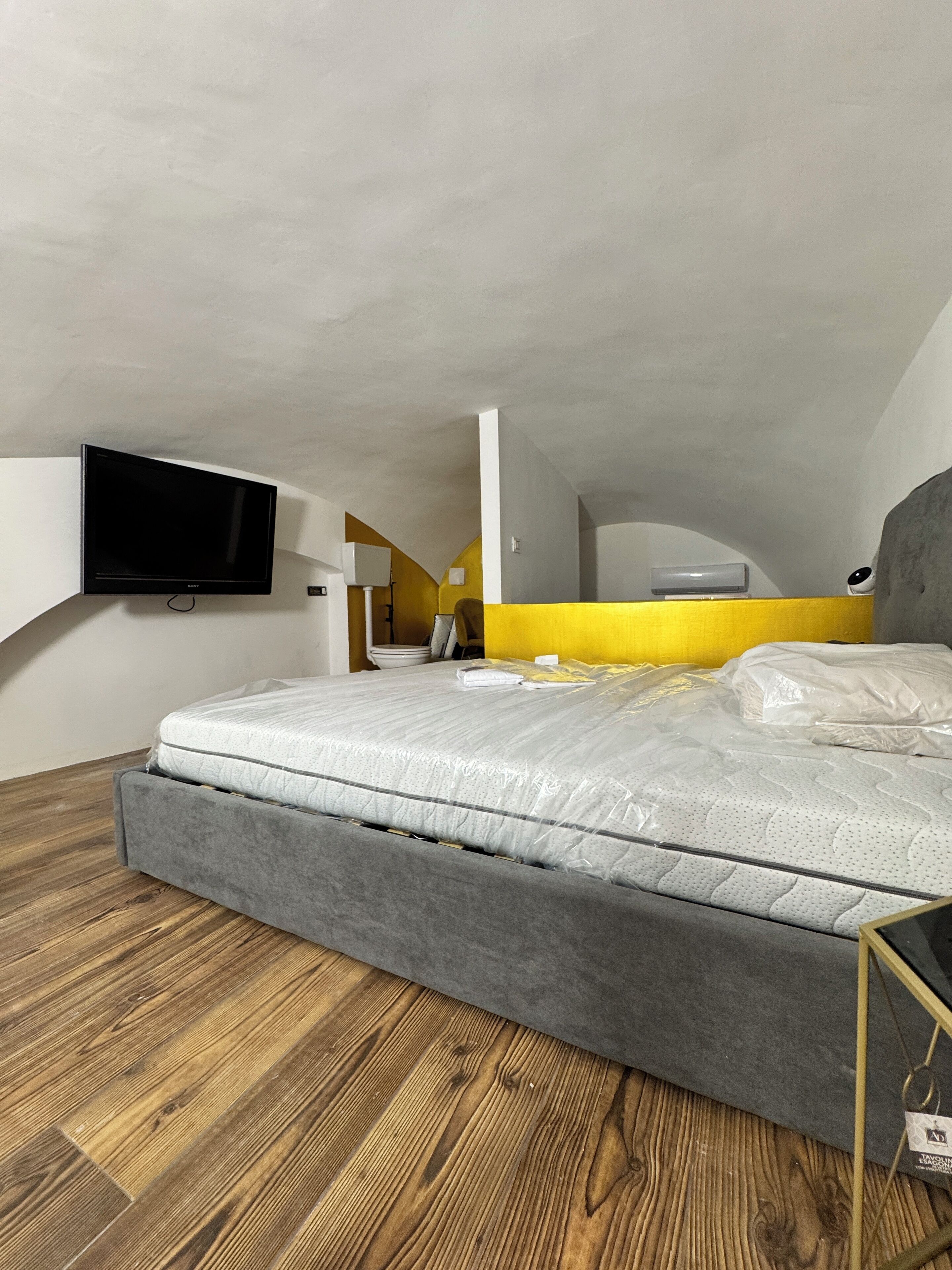 Quarto com cama, piso de madeira, televisão na parede e uma parede amarela como detalhe.