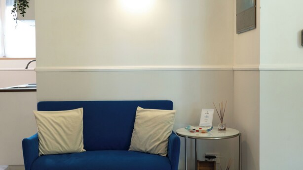 Un canapé bleu avec deux coussins, une petite table ronde sur laquelle se trouvent un bocal en verre et une boîte, et un luminaire fixé au mur.