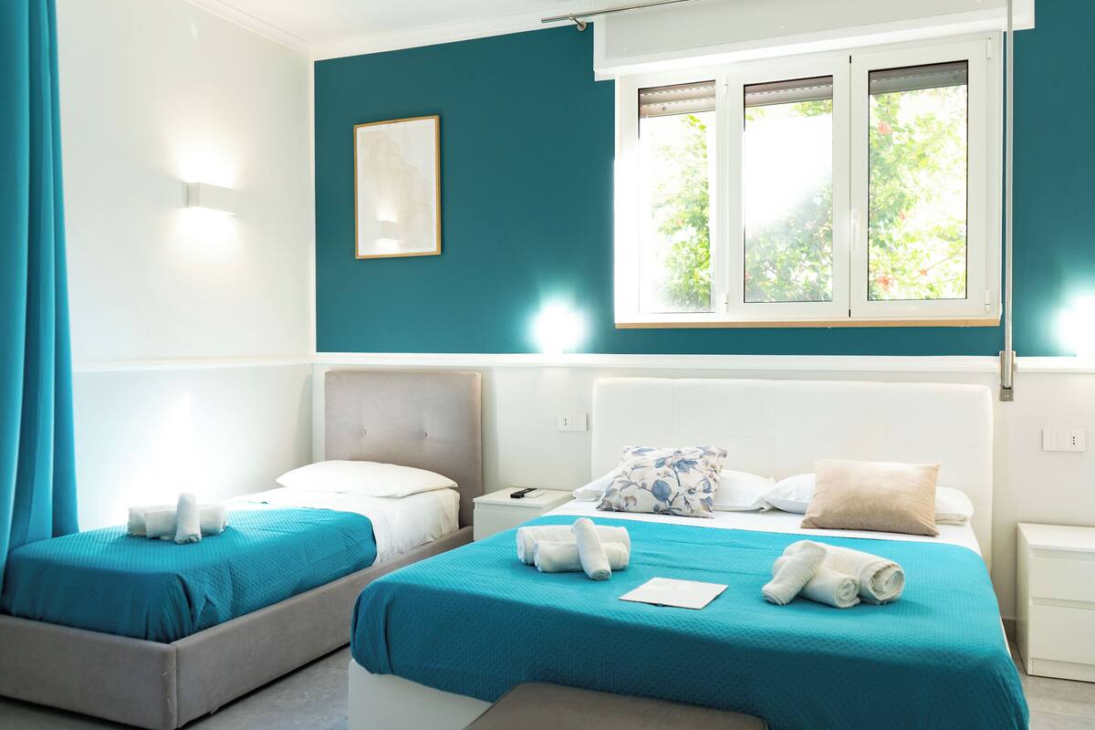 A hotel room with two beds, teal walls, a window with blinds, a bedside table, and a nightstand.