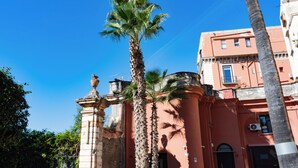 Un bâtiment devant lequel se trouve un palmier, une statue sur un piédestal et un ciel d’un bleu limpide.