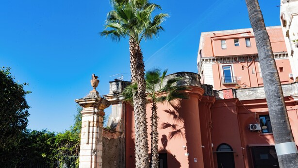 Un bâtiment devant lequel se trouve un palmier, une statue sur un piédestal et un ciel d’un bleu limpide.