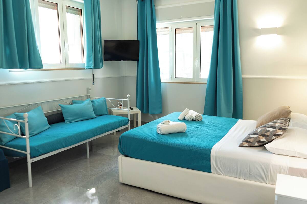 A hotel room with a bed, a sofa bed, a TV mounted on the wall, and a window with curtains.