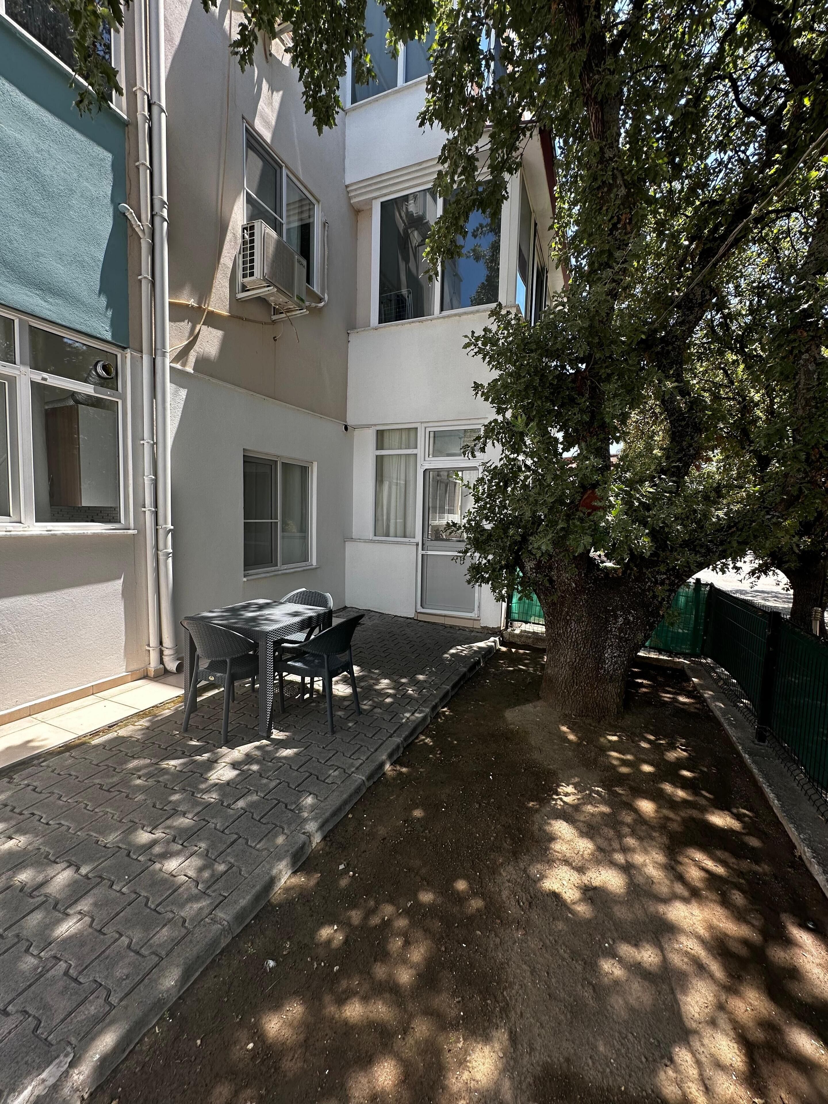 A courtyard with a table and chairs, surrounded by buildings with multiple windows and a tree providing shade.