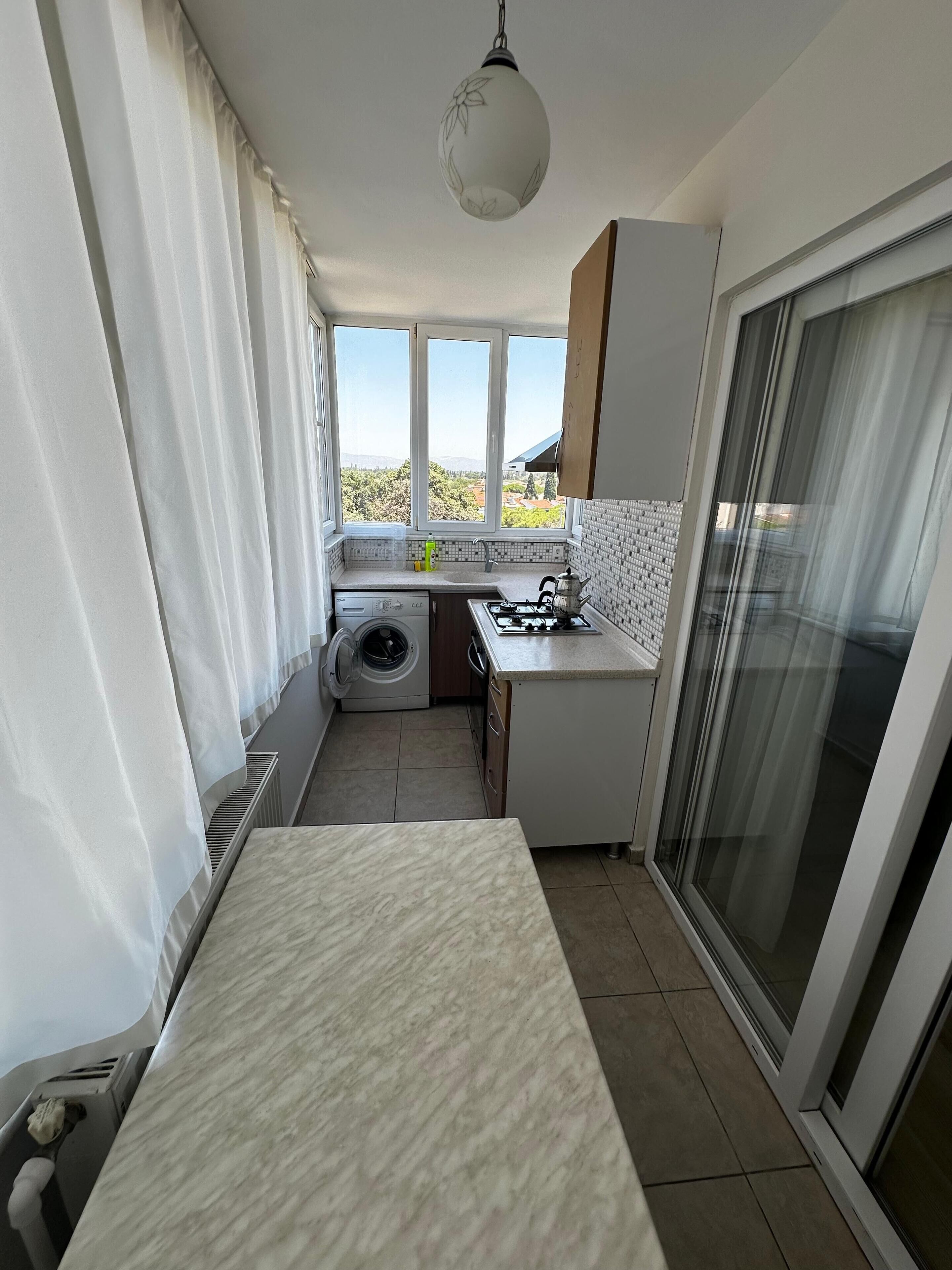 A compact kitchen with a washing machine, a sink, and a stove. There is a large window providing a view of the outdoors.