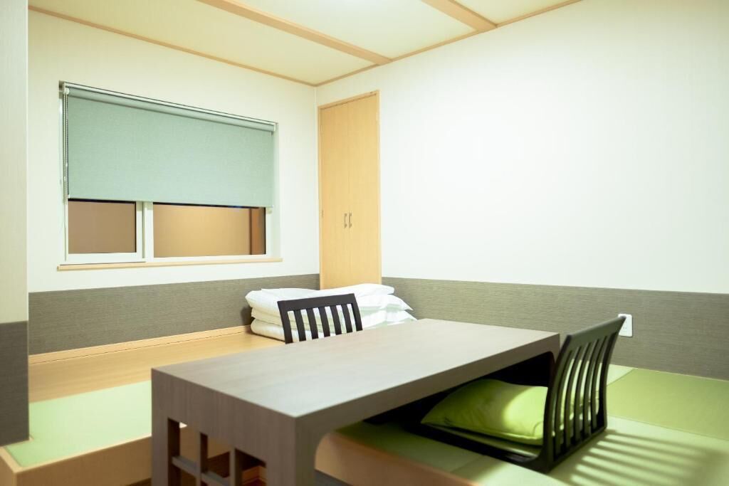 Una habitación con una cama, una mesa y una silla. Se ve una ventana con persiana, una puerta y una pared con interruptor de luz.