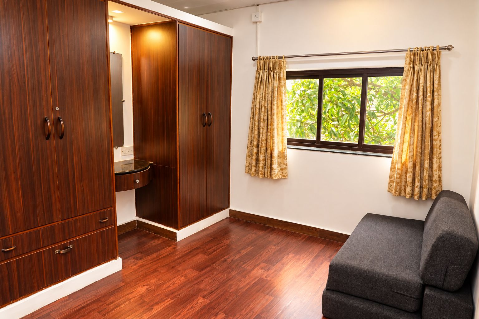 A room with a wooden wardrobe, a small sink, a window with curtains, and a grey sofa.