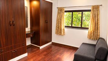 A room with a wooden wardrobe, a small sink, a window with curtains, and a grey sofa.