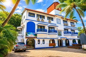 Un hotel a più piani con balconi, un'auto parcheggiata davanti e palme circostanti.