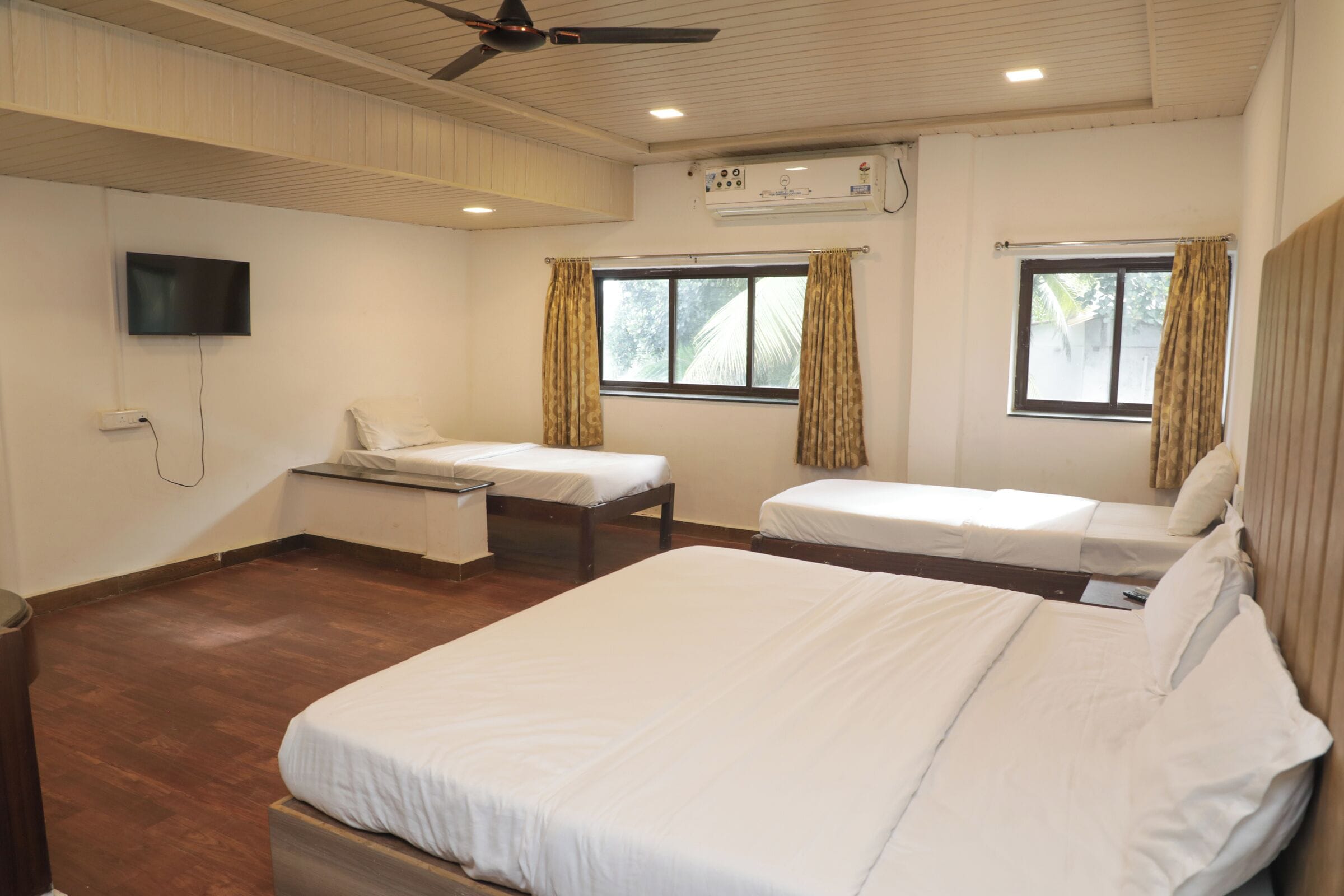 Habitación de hotel con dos camas individuales, ventilador de techo, unidad de aire acondicionado, televisor montado en la pared y ventana con cortinas.