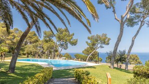 Zona de piscina con terraza de madera, rodeada de árboles y vista al mar.