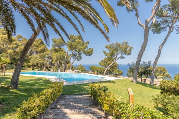 A pool area with a wooden deck, surrounded by trees and a view of the sea.