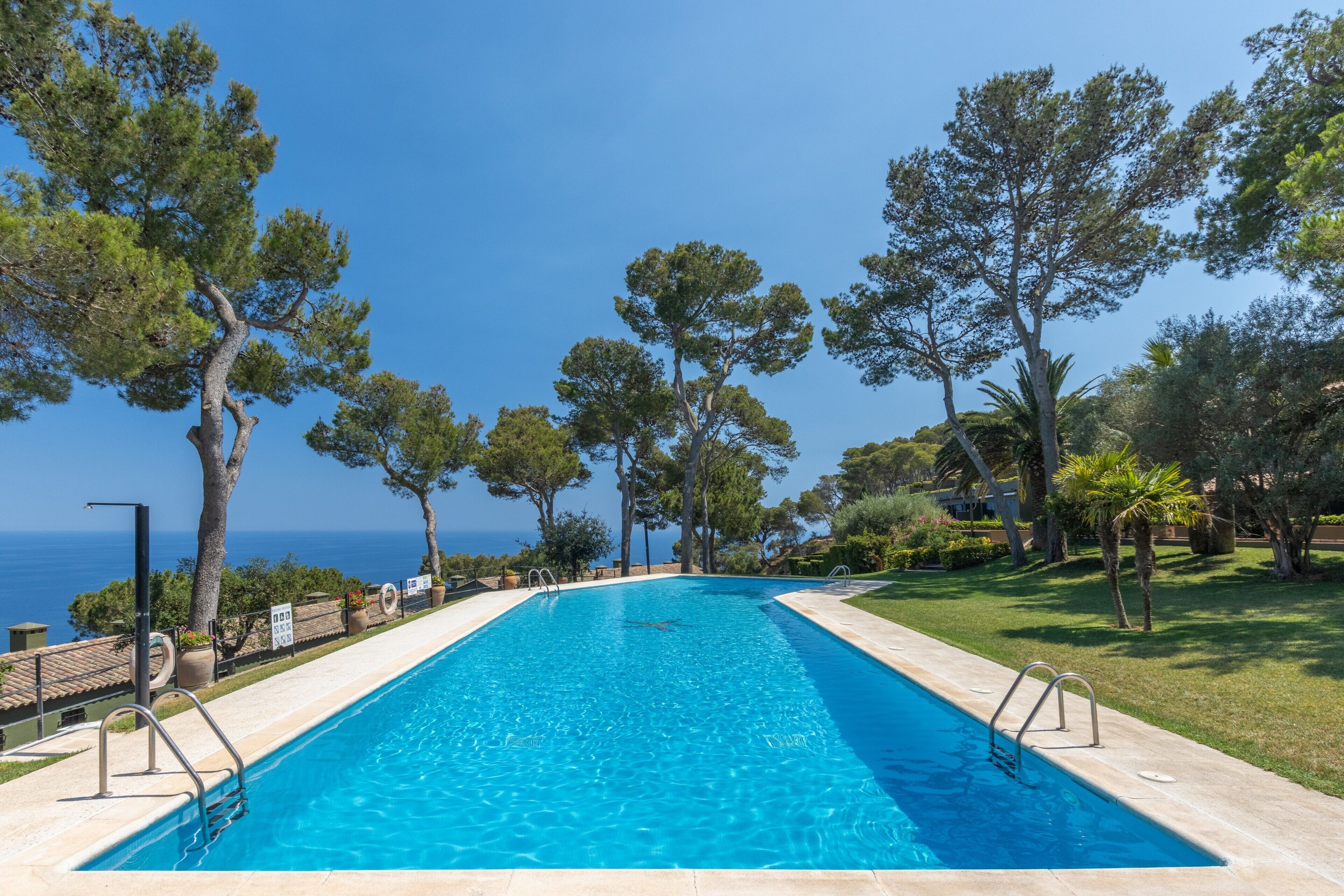 Una piscina rectangular con agua cristalina y azul, rodeada por una solana empedrada y barandillas de metal.