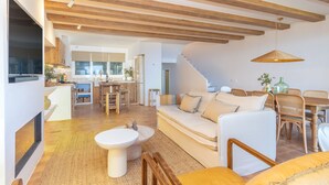 Una sala de estar moderna con sofá, mesa de comedor y sillas, vigas de madera en el techo, y una cocina al fondo.