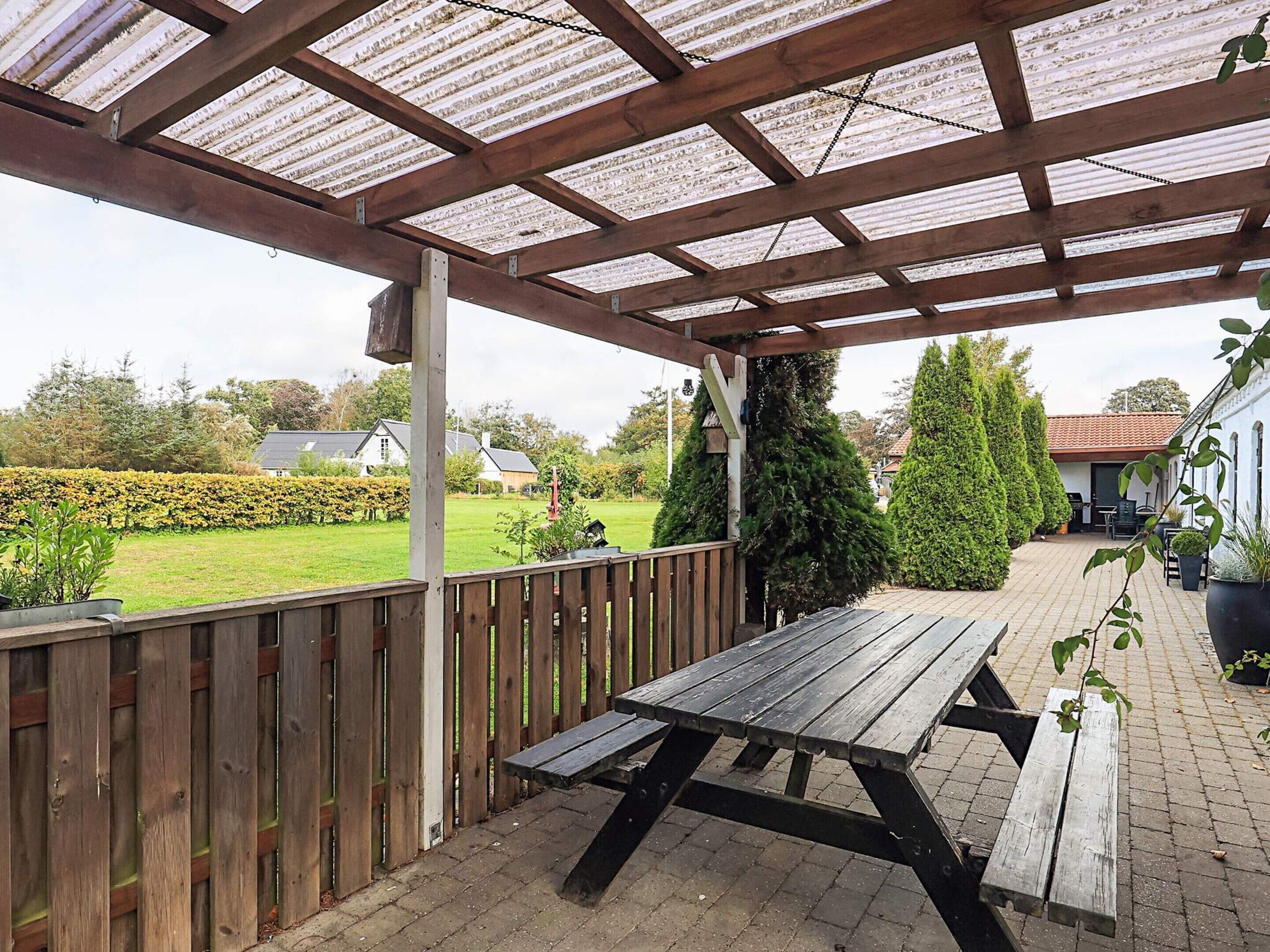 Un patio techado con una mesa y bancos de madera, una valla de madera, y vistas a un jardín y casas al fondo.