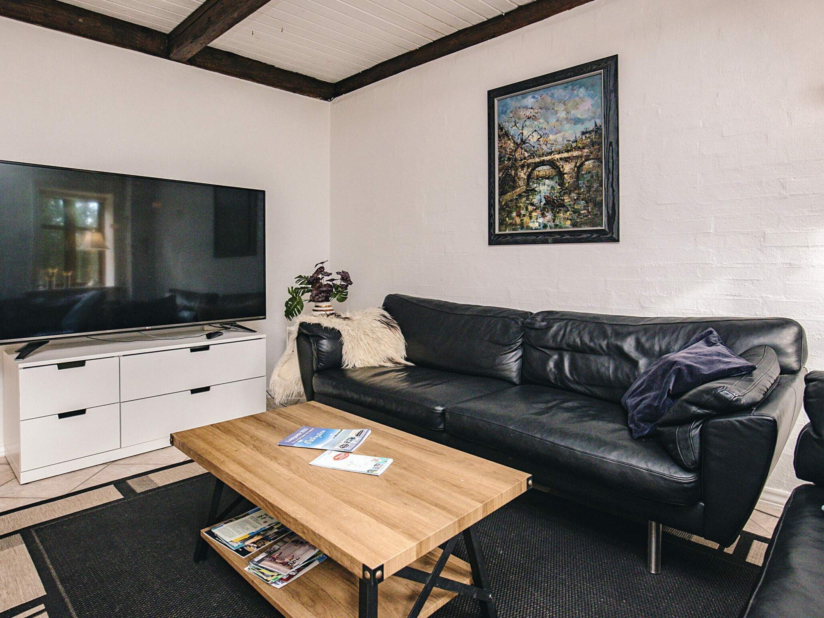 A modern living room with a black leather sofa, a wooden coffee table, a flat-screen TV, a white cabinet, and a framed painting on the wall.
