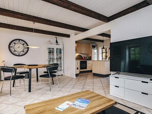 A modern kitchen with white cabinets, a dining area with a wooden table and chairs, a large wall clock, and a television.