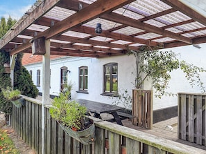 A covered patio area with a wooden table and benches, potted plants, and a white building with windows in the background. - 4 Star Holiday Home in Asaa (Asaa)