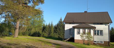 A two-story house with a brown roof and white walls, surrounded by trees and a grassy area.