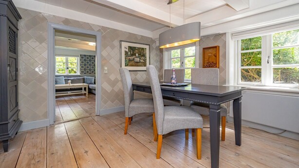 A dining area with a wooden table and chairs, a window with a view of greenery, and a wall with a diamond-patterned tile design. - Kapitänshuis - Captain's house on Sylt with garden, terrace and comfortable (Sylt)