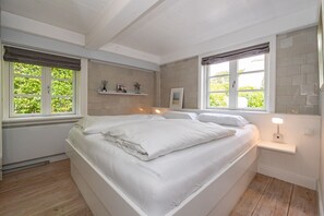 Quarto com cama grande, duas janelas com cortinas, mesa de cabeceira com abajur e prateleira com objetos decorativos.