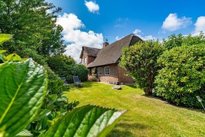 Een stenen huis met rieten dak, omgeven door weelderige begroeiing en een verzorgd gazon.