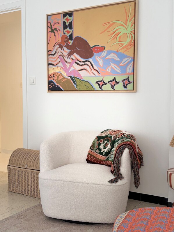 A white armchair with a colorful throw blanket, a wicker basket, and a framed abstract painting on the wall. - Let's Go Koksijde (Koksijde)
