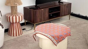 Una sala de estar con televisor sobre un soporte de madera, una lámpara a rayas y un otomano estampado.