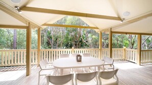 A covered outdoor area with a table and chairs, surrounded by a wooden deck and railing, overlooking a forested area.
