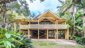 A two-story wooden house with a balcony, surrounded by tropical vegetation.