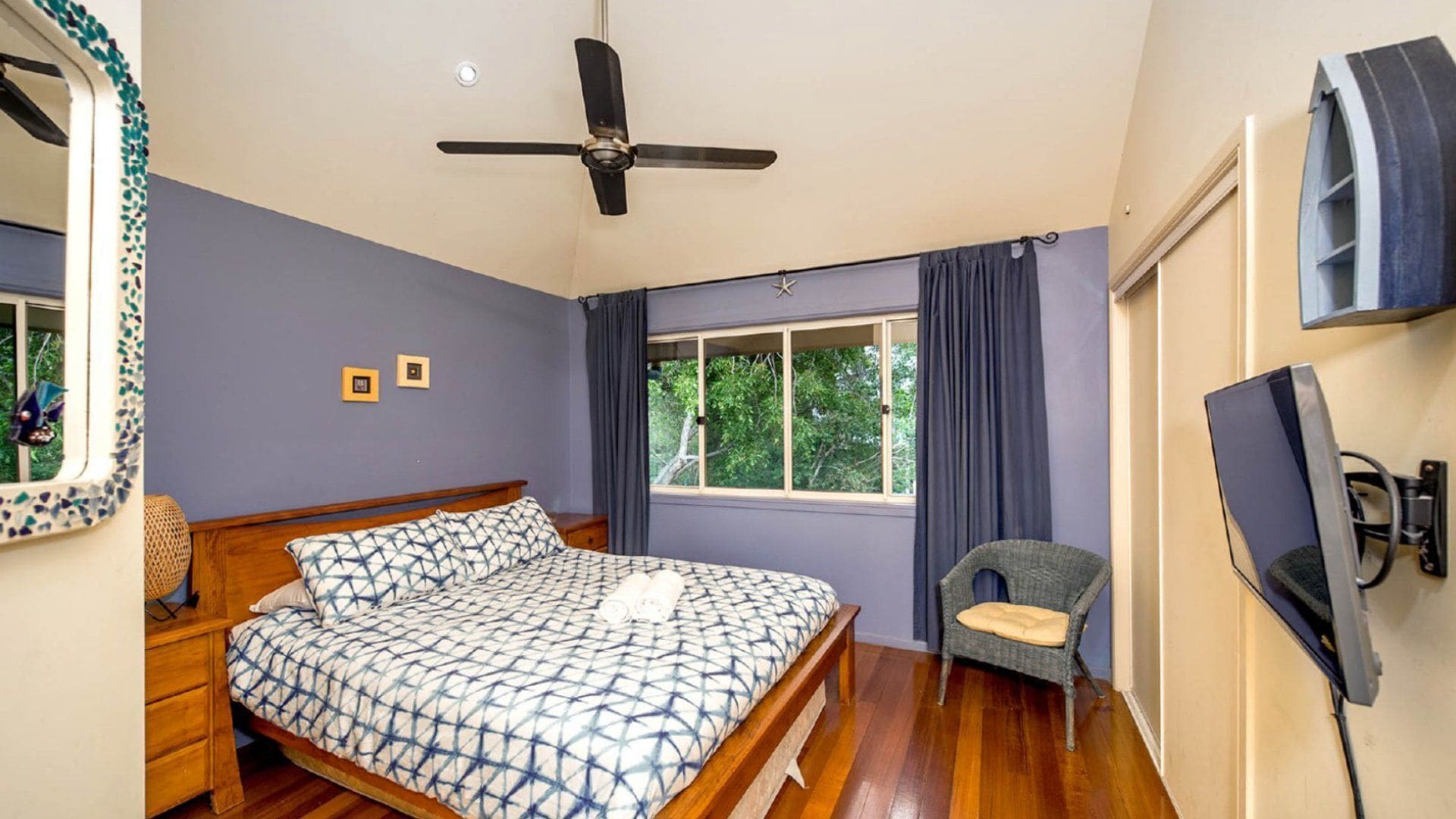 A bedroom with a bed, a chair, a television, a ceiling fan, and a window with curtains.