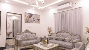 Un salon bien éclairé, avec deux canapés richement décorés, une table basse sur laquelle est posé un panneau, et un grand miroir accroché au mur.