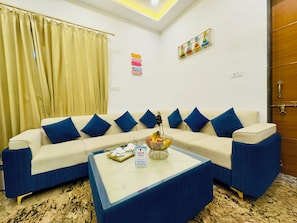 Una sala de estar con un sofá color beige, cojines azules, una mesa de centro con fruteros y una puerta con picaporte de latón.