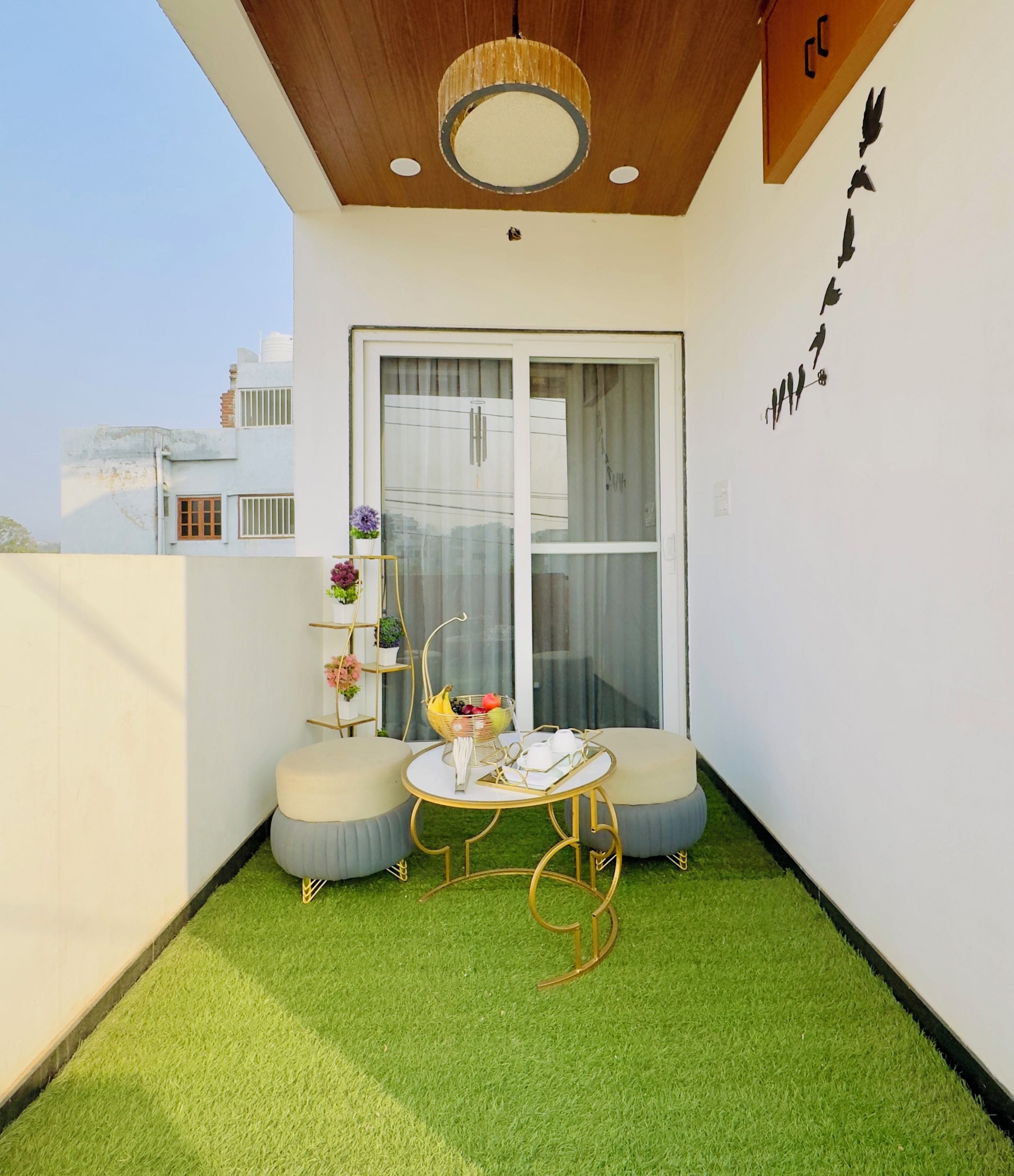 A balcony with artificial grass, a round table, two cushioned seating, a hanging light fixture, and a window with curtains.