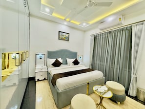 A hotel room with a bed, bedside tables, a ceiling fan, an air conditioning unit, a window with curtains, and a small table with a tray. - Sagar Villa Udaipur (Udaipur)