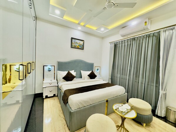 Habitación de hotel con cama, mesitas de noche, ventilador de techo, equipo de aire acondicionado, ventana con cortinas y una pequeña mesa con bandeja.