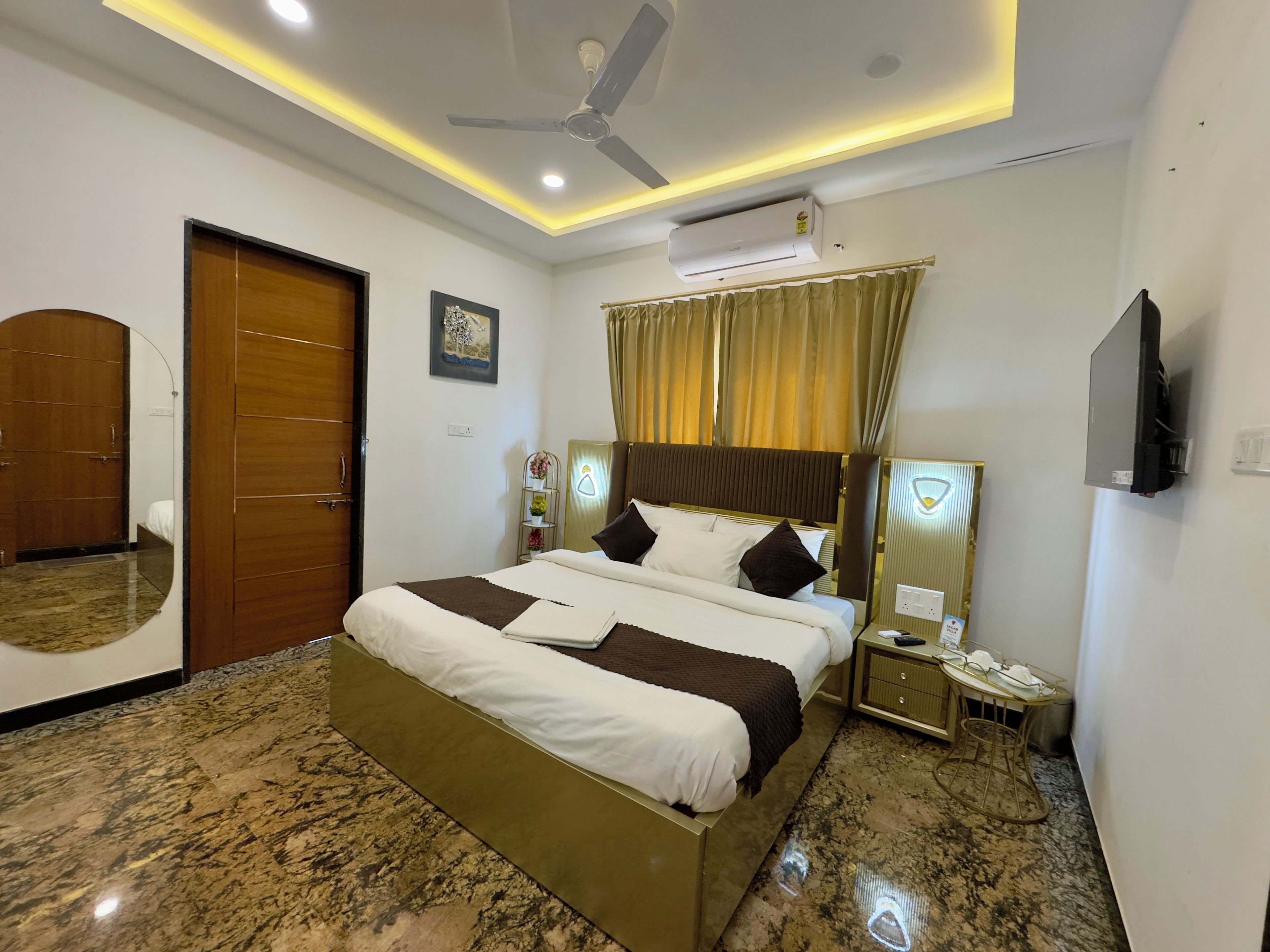 A hotel room with a bed, a ceiling fan, an air conditioning unit, a bedside table with a lamp, and a wall-mounted television.
