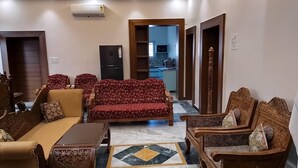 A living room with sofas, a coffee table, and a TV. There is a kitchen visible through an open doorway. - Manoram Hospitality (Navi Mumbai)
