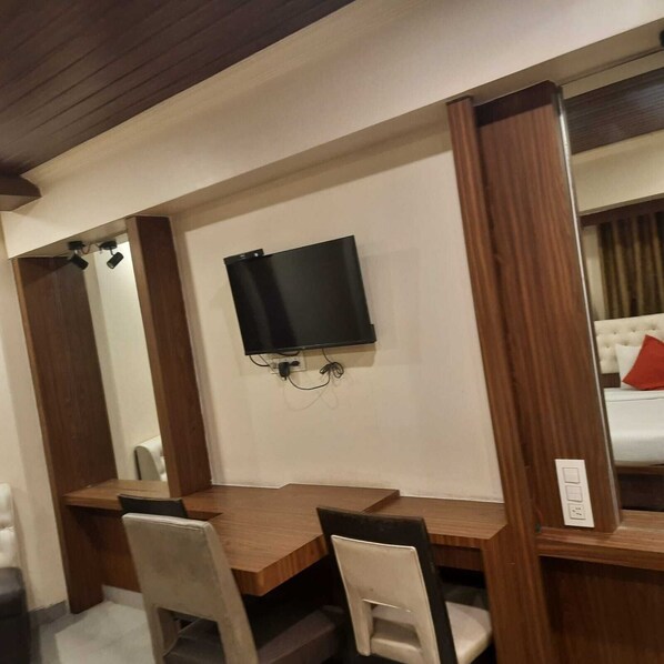 A hotel room with a wooden desk, two chairs, a flat-screen TV mounted on the wall, and a bed with white bedding and a red pillow.