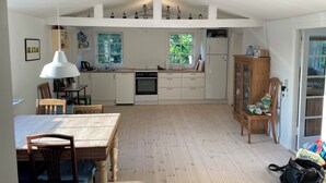 A compact kitchen with white cabinets, a built-in oven, and a refrigerator. There is a dining table with chairs, a wooden cabinet, and a window providing natural light.