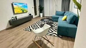 A modern living room with a blue sofa, a white rocking chair, a black and white patterned rug, and a flat-screen TV mounted on the wall.